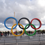 a sculpture of the five interlocked multicolor rings of the olympic logo stands in front of the metal latticework Eiffel Tower in PAris